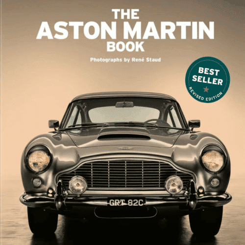 Lyxig coffee table-bok med titeln Aston Martin – Tankad och redo att köra (110-årsjubileumsutgåva). Boken ligger uppslagen på ett bord och visar högupplösta studiobilder av klassiska och moderna Aston Martin-bilar, fotograferade av René Staud. Den eleganta inbundna volymen har ett stilrent omslag och symboliserar 110 år av brittisk design, ingenjörskonst och bilhistoria.